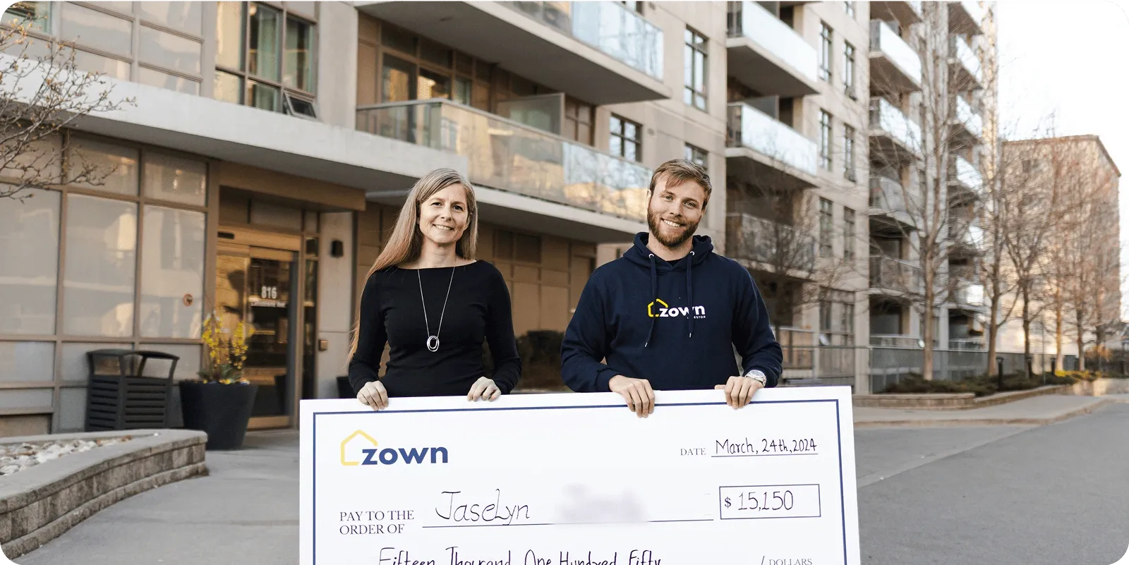 Homeowners standing in front of a condo building holding a large Zown cheque for down payment assistance, representing real clients who received financial support and successfully purchased their home.